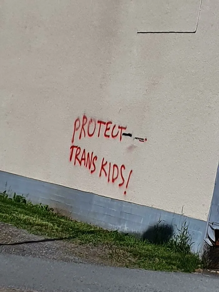 Graffiti vandalism on public property with red spray paint message protecting trans kids on a beige wall.