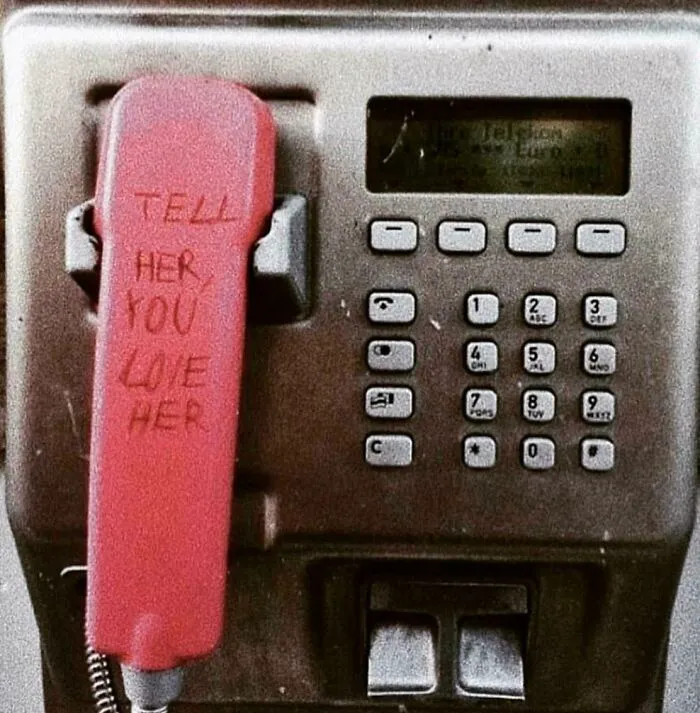 Old public payphone with vandalized red handset engraved with message telling someone to confess love.
