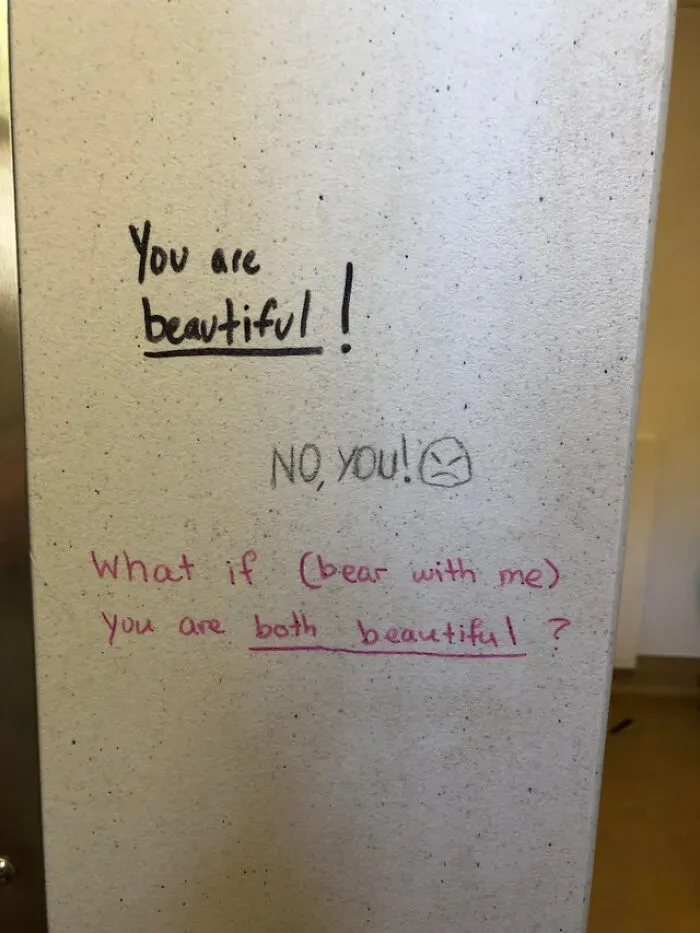 Positive and playful public property vandalism featuring compliments and a small angry face drawn on a wall.