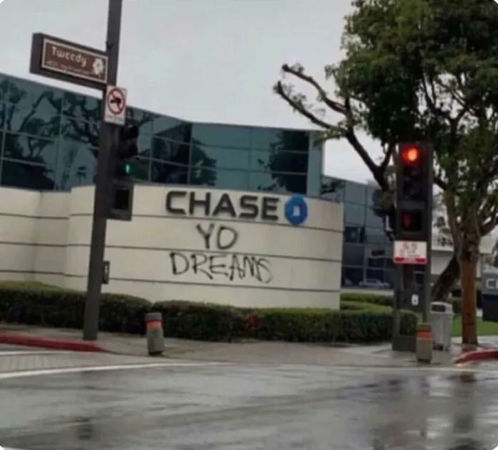 Chase bank building with graffiti vandalism reading "YO DREAMS" on public property near traffic lights and trees.