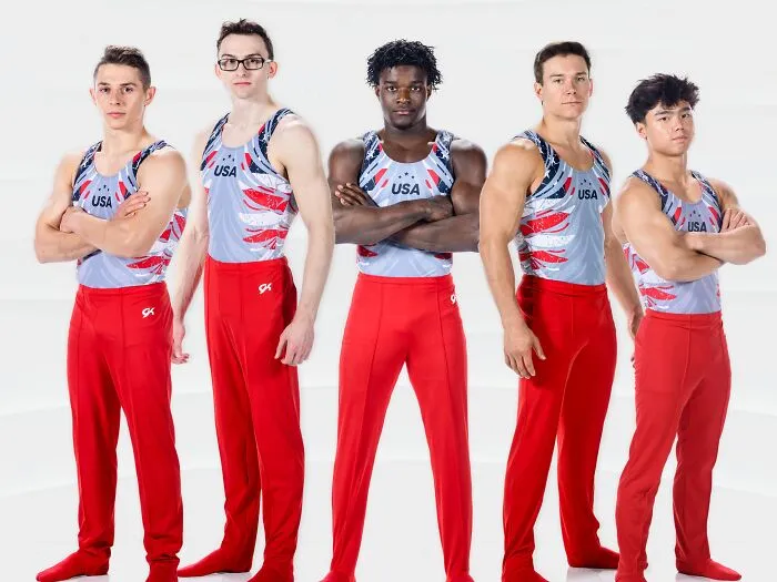 Five male athletes wearing USA gymnastics uniforms with red pants and sleeveless tops standing with arms crossed.
