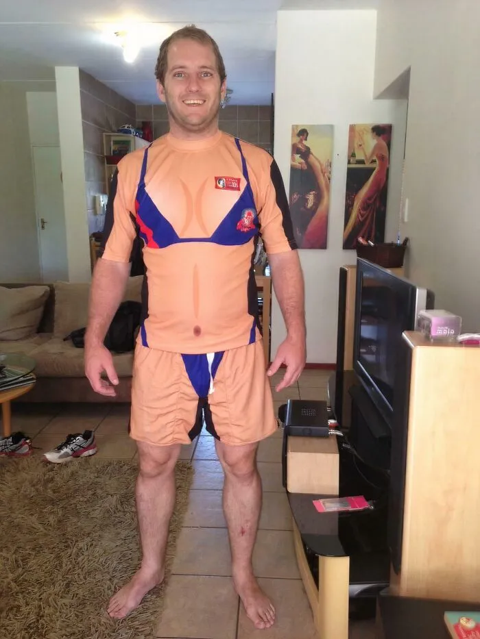 Man wearing a funny printed uniform resembling a bikini, standing barefoot in a living room with casual decor.