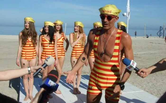Group wearing matching funny striped swimsuits and hats on the beach during a humorous uniform event with media coverage.