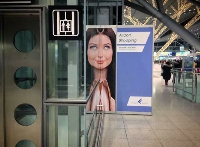 Advertising placement fail showing a distorted face on an airport shopping poster next to restroom sign causing humor.