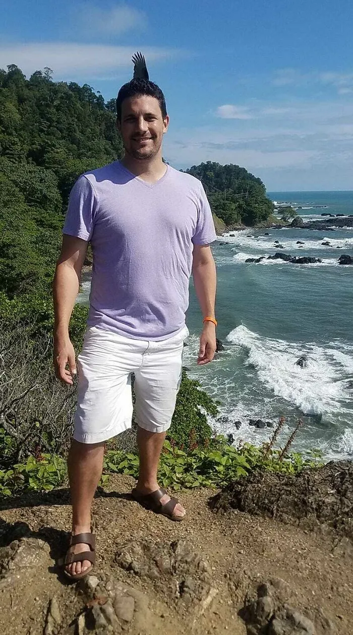 Hombre sonriente en la playa con un pájaro posado que crea una foto mejor de lo esperado con naturaleza al fondo.
