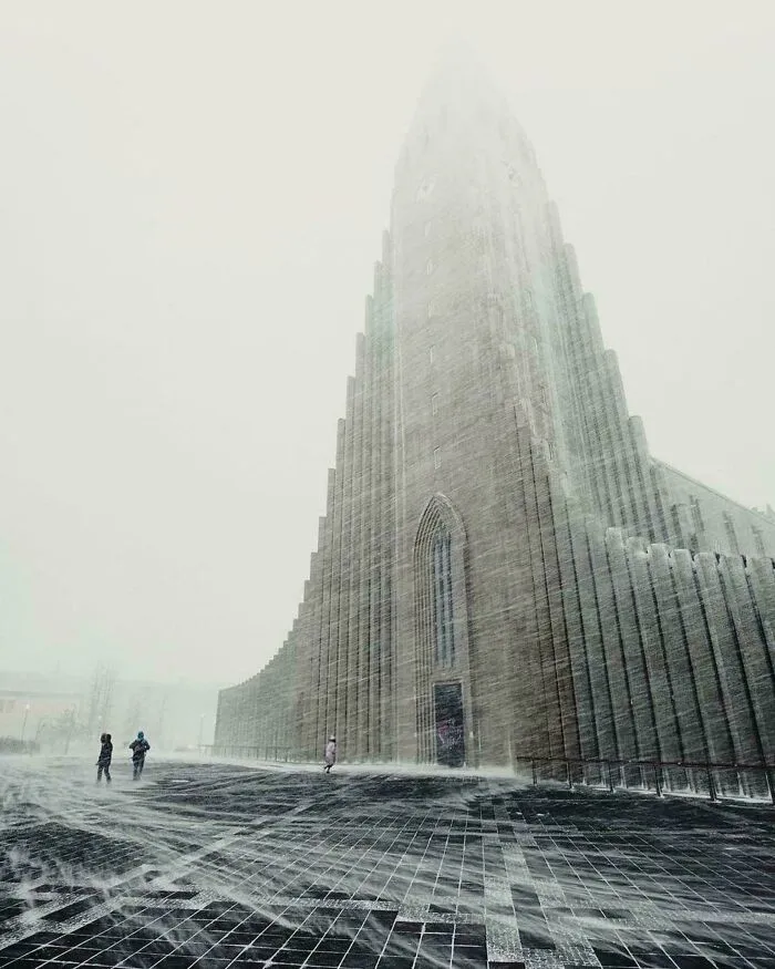 Three people brave the snow in front of an extremely large and stark church, an eerie and majestic structure.