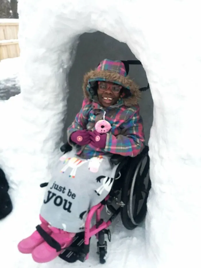 Child in a wheelchair smiling inside a snow fort, wearing colorful winter clothes in a heartwarming story setting.