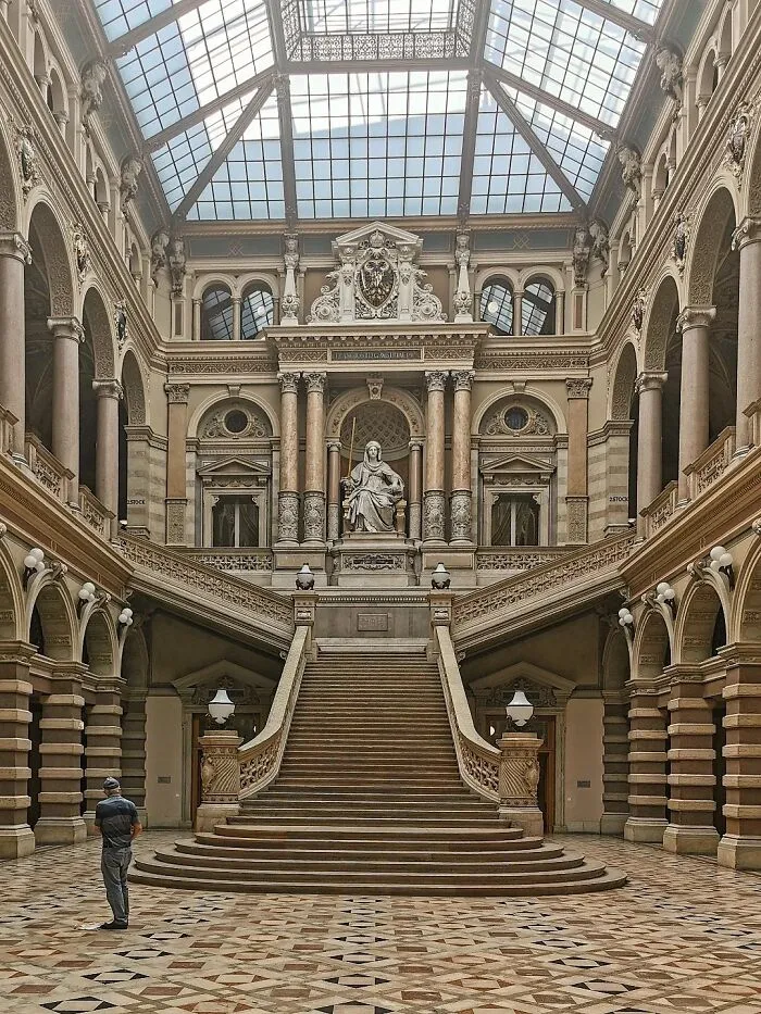 Grand interior architecture with a sweeping double staircase leading to a statue, surrounded by columns and ornate details under a glass ceiling.