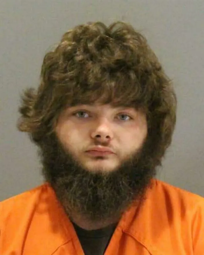 A mugshot of a man with a thick, curly brown beard and matching hair, showing an unfortunate haircut.