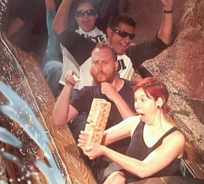 Personas con gran sentido del humor jugando Jenga en parque de diversiones, capturadas con expresiones divertidas.