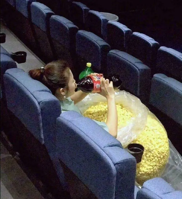 Woman drinking soda while sitting with an enormous bag of popcorn in a movie theater, funny and bizarre image combining the holy and cursed.