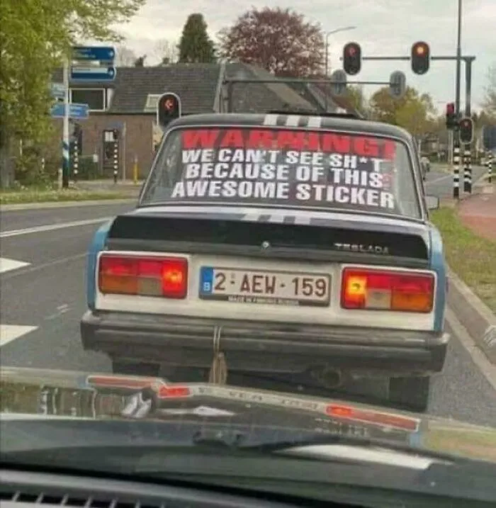 Old car with a funny and bizarre sticker covering the rear window, blending humor with a cursed and holy vibe on the road.