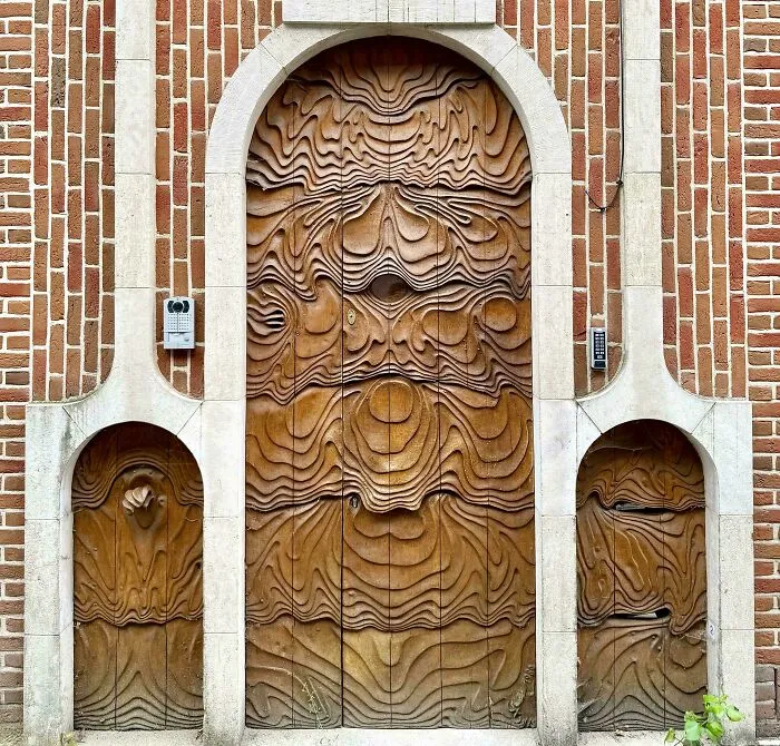 An elaborate wooden door with a wavy, textured design, flanked by two smaller arched doors, all set within a brick facade.