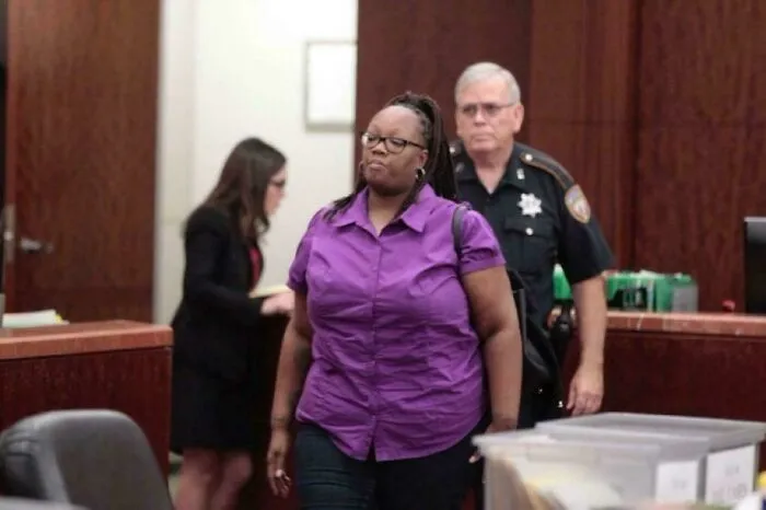 Woman in purple shirt escorted by police officer in courtroom, with legal staff working in the background, still remains undiscovered