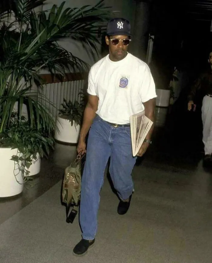 Man wearing a New York Yankees cap, sunglasses, white t-shirt, and jeans, holding a bag and folded papers, 90s style.