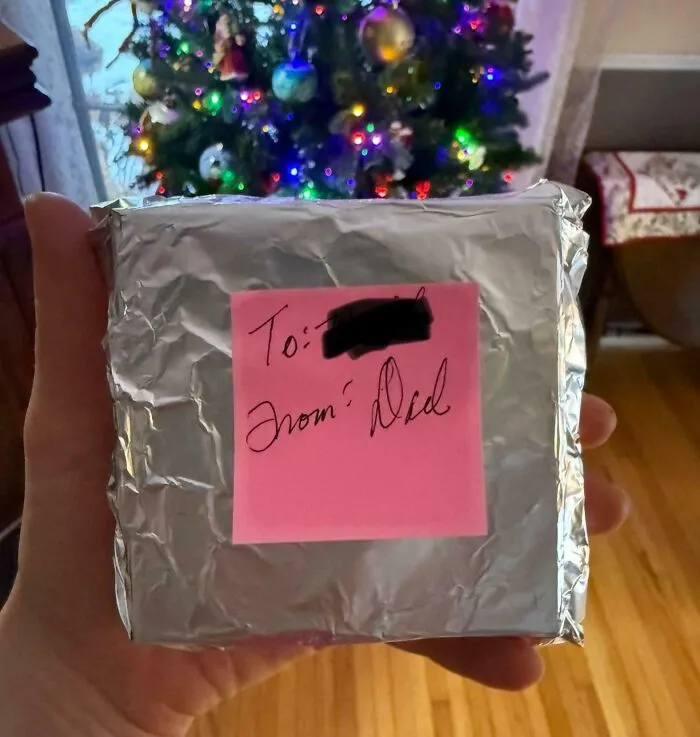 Hand holding a crudely wrapped gift with a pink note labeled from Dad, illustrating funny photos of moms and dads differences.