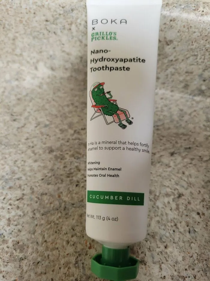 Tube of cucumber dill flavored nano-hydroxyapatite toothpaste with a quirky pickle cartoon on a speckled countertop.