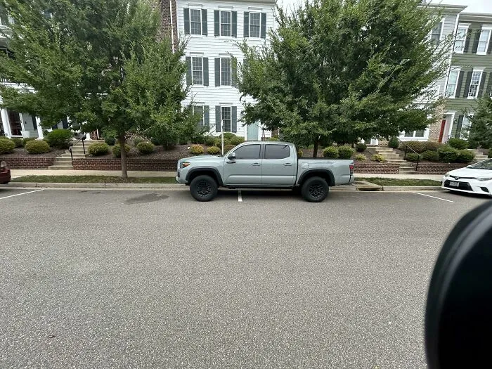 Camioneta gris estacionada en dos espacios en calle residencial con casas blancas y árboles al fondo. Incidentes expuestos.