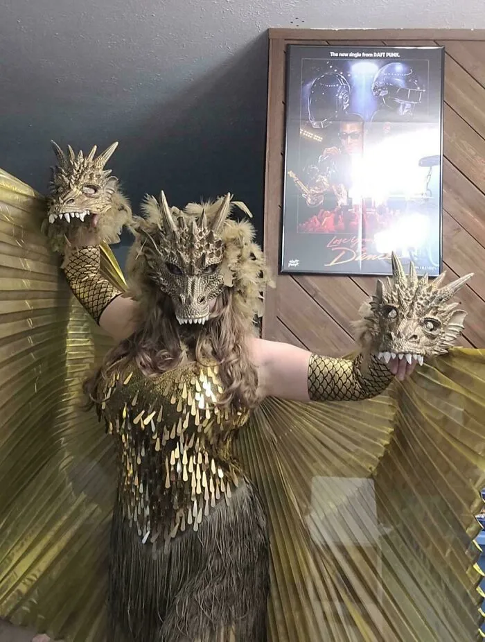 Person in a detailed dragon costume with gold scales and wings, holding two dragon head props for Halloween costumes.