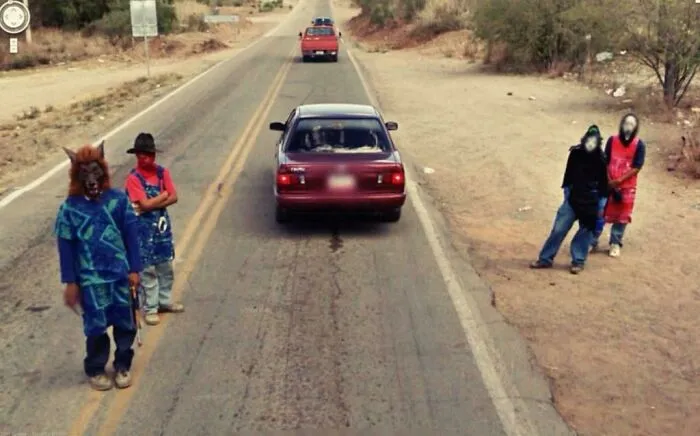 Four people wearing strange and unexplained costumes stand by a rural road next to a red car.