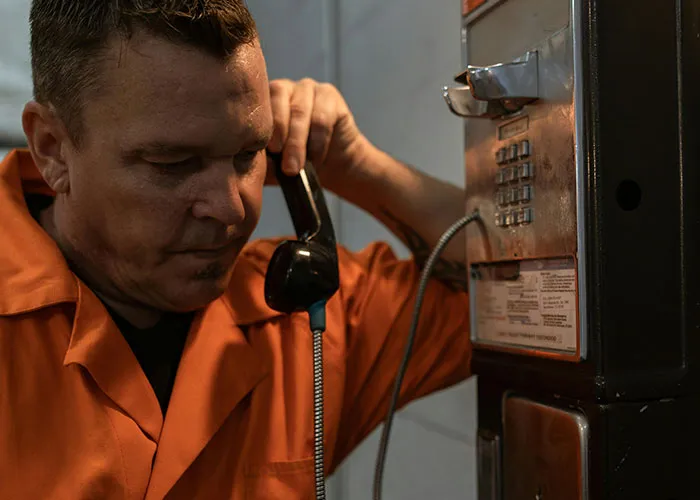 Man in an orange jumpsuit using a payphone looking serious, representing criminal fails and humorous mistakes.