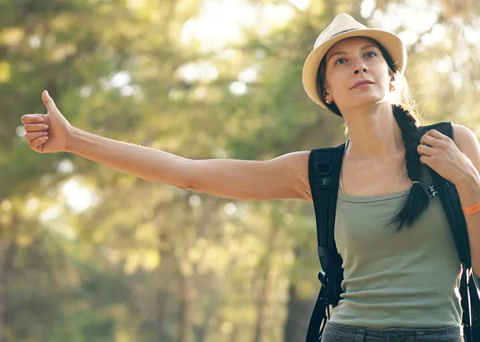 Young woman with a hat and backpack hitchhiking on a forest road, evoking terrifying true stories vibe.