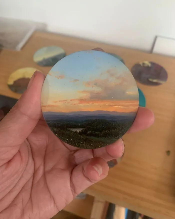 Hand holding a detailed painting by Dina Brodsky of a natural landscape with a sunset and distant mountains.