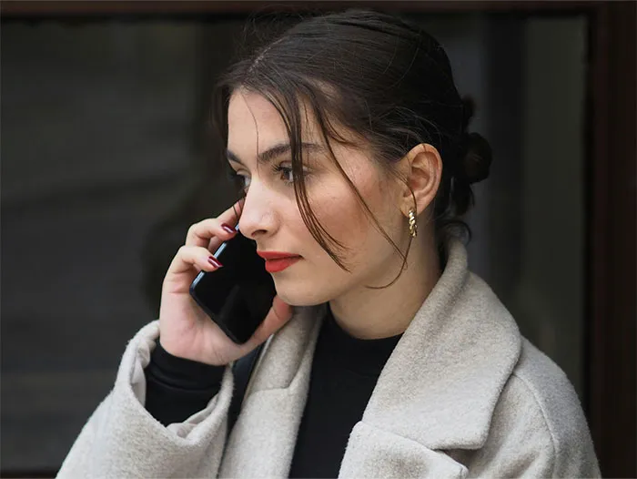 Young woman in a beige coat talking on a phone, reflecting on bizarre and paranormal experiences she can't explain.