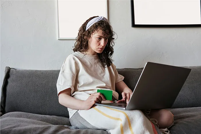 Young woman with curly hair and headband using laptop on sofa, focused on researching bizarre and paranormal experiences online.