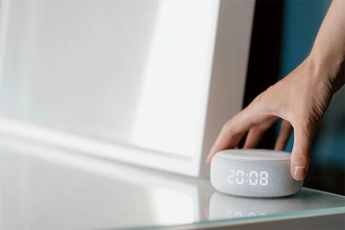 Hand reaching to press a smart speaker clock on a glass table, evoking strange and paranormal experiences concept.