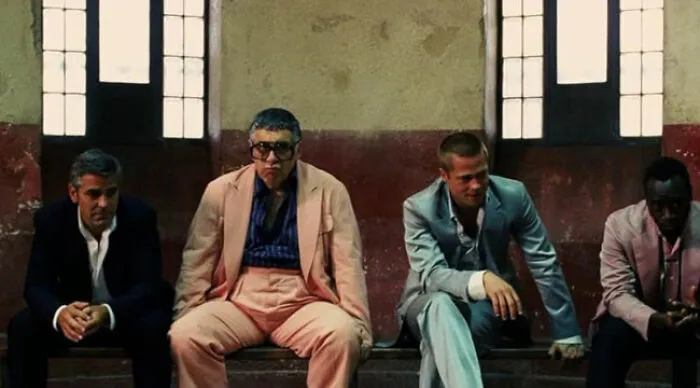 Four men dressed in suits sitting on a bench in a dimly lit room, representing beloved movie tropes.