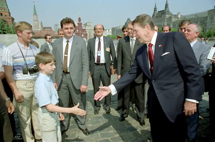 Young boy reaching to shake hands with a suited man amid onlookers in an outdoor historical setting, photo mysteries revealed.