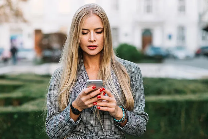 Young woman with long blonde hair in checkered jacket looking at phone outdoors, reflecting on catfish stories and online DMs.