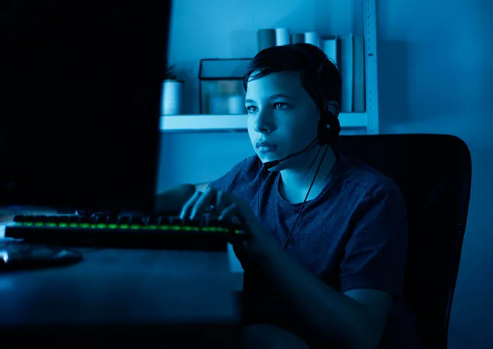 Teen boy wearing headset and using computer in dark room, depicting catfish stories and online deception risks.