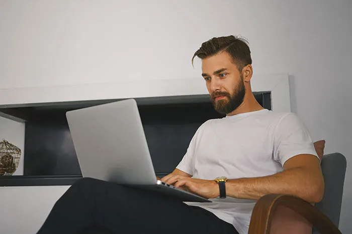 Man with beard wearing white shirt, sitting with laptop, reading catfish stories that question every DM he received.