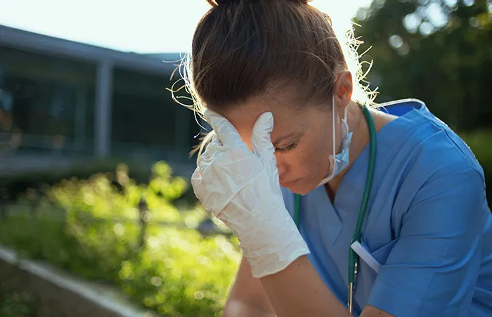 Healthcare worker in blue scrubs and gloves resting outdoors, showing stress and exhaustion after a long shift.