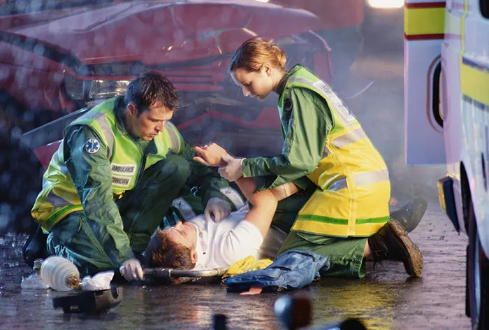 Healthcare workers attending to an injured man on the ground after a car accident, providing urgent medical care.
