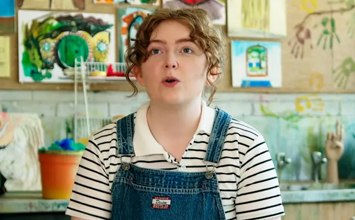 Young woman in striped shirt and denim overalls speaking about experiences with celebrities before fame in art-filled room