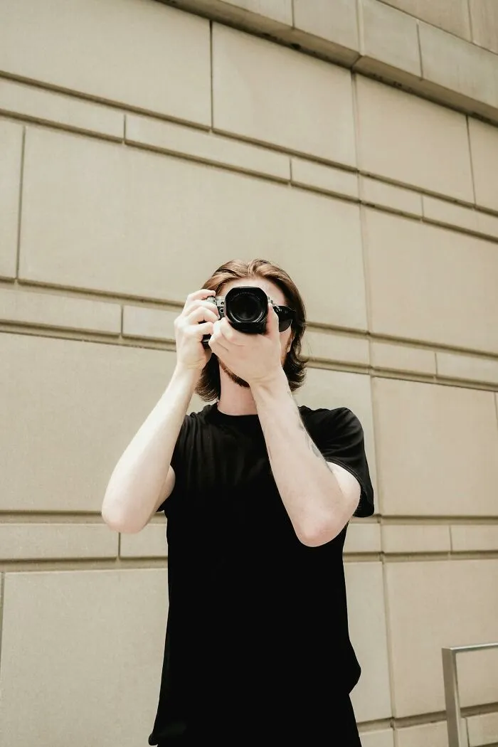 Young man holding a camera taking a photo against a beige wall, illustrating people who were the villain in someone else's story.