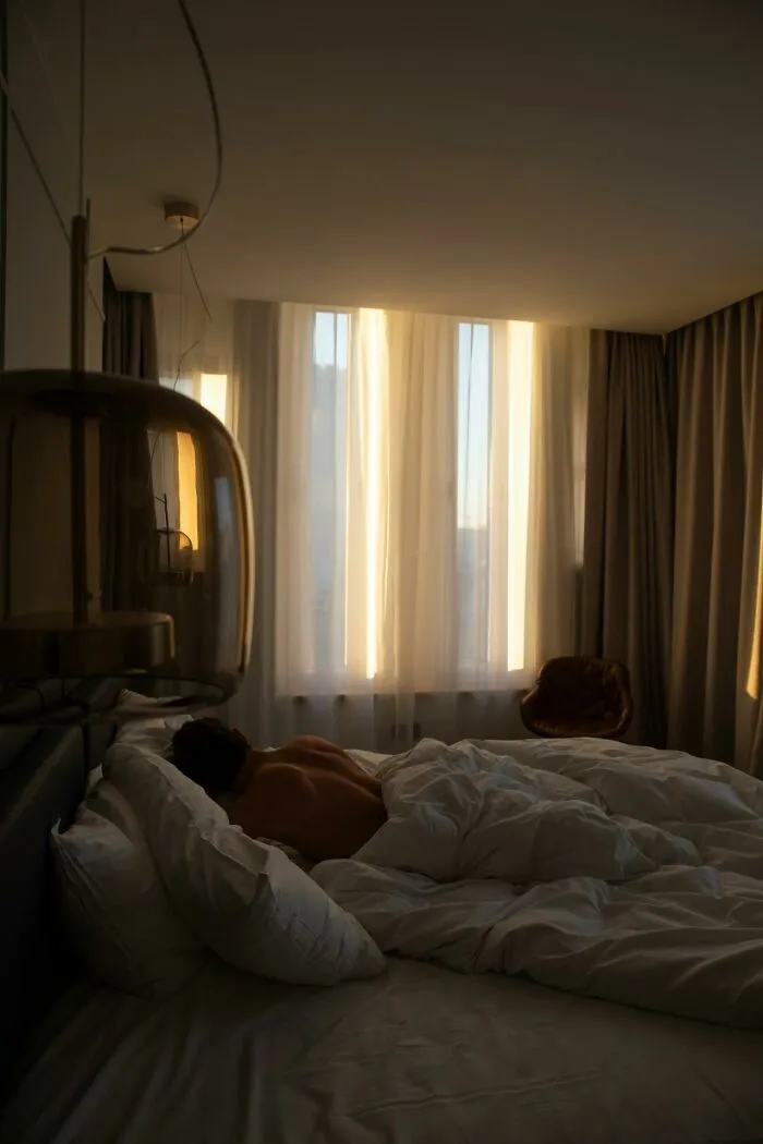 Person lying in bed in softly lit room with curtains during sunrise, reflecting on consequences of dumb decisions.