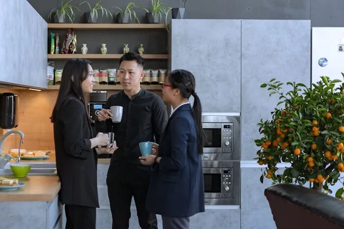 Three coworkers talking and drinking coffee in a modern kitchen during a break discussing workplace mistakes and fired employees.