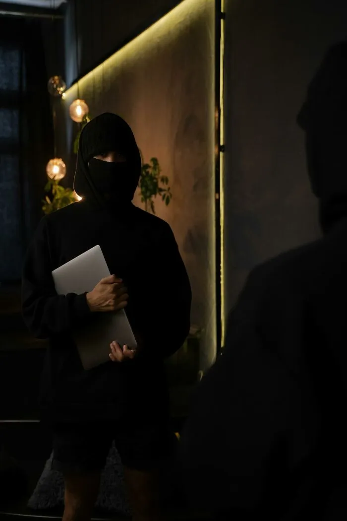 Person in dark hoodie and face mask holding laptop in dimly lit room, reflecting themes of getting fired for dumb mistakes