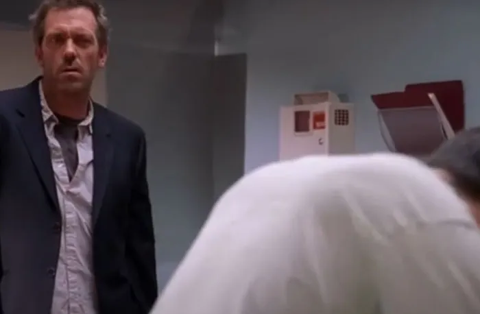 Scene from House M.D. showing Dr. House observing another doctor in a clinical setting with medical equipment nearby.