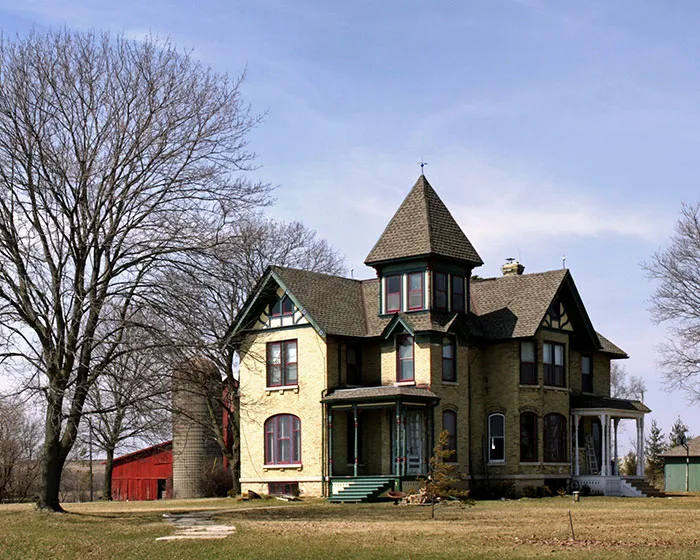 Old spooky Victorian house with bare trees and a red barn, evoking scary real-life stories and eerie atmosphere.
