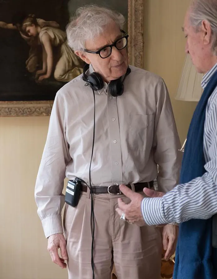 Older male star wearing glasses and headphones, dressed in beige, engaged in conversation about male stars' actual height.