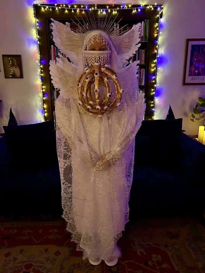 Person in a genius Halloween costume wearing intricate white lace with wings and a crown adorned with eyeballs and lights.