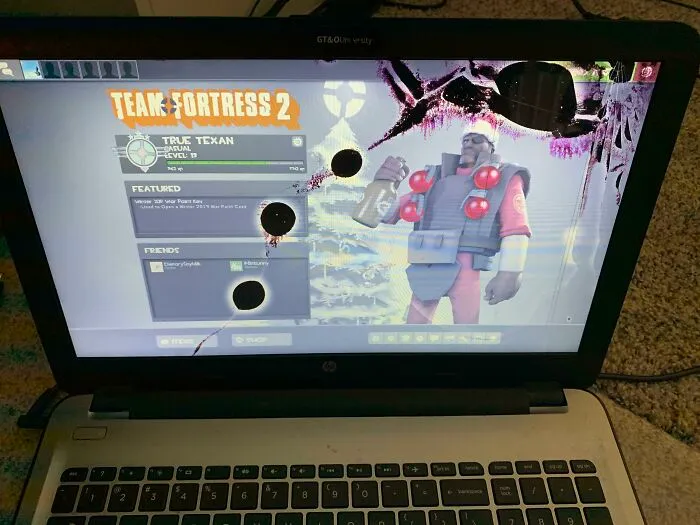 Laptop screen heavily cracked and damaged, showing a Team Fortress 2 game while handling electronics the wrong way.