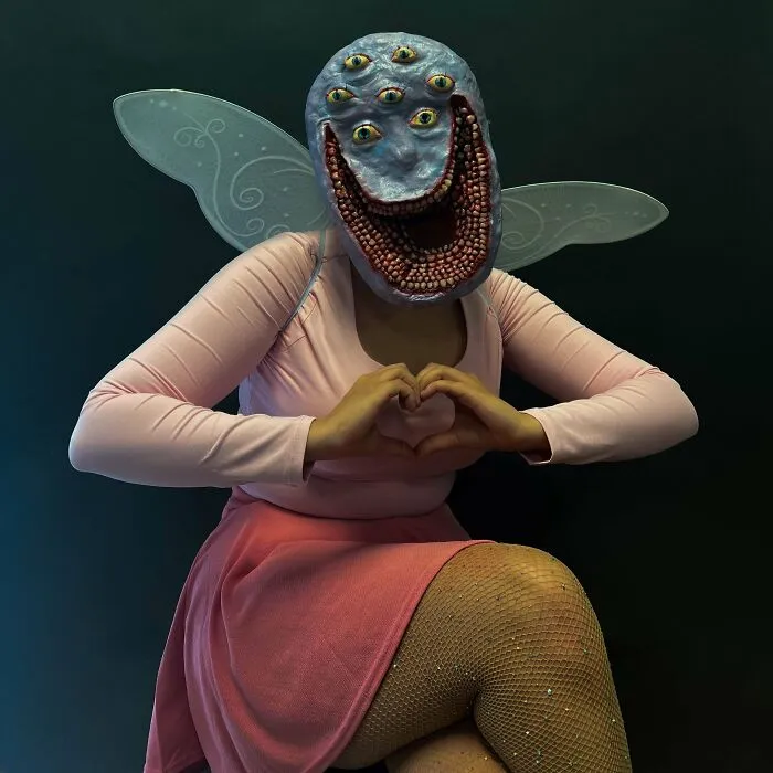 Person wearing a genius Halloween costume with multiple eyes, wings, and a pink outfit posing with hands in a heart shape.