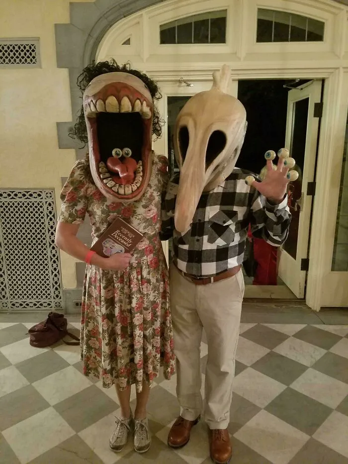 Two people in genius Halloween costumes, one with a large mouth mask holding a book, the other with a bird-like mask and eyeball fingers.
