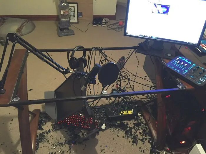 Messy electronics setup with broken glass, tangled cables, and scattered components under a desk and monitor.